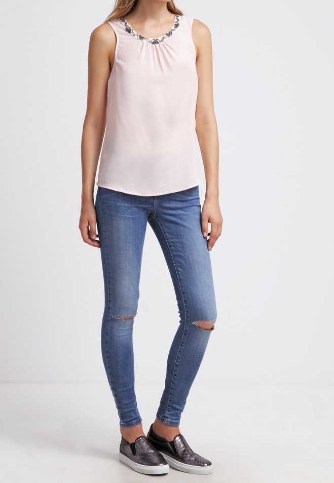 Femme portant un haut rose clair sans manches avec un col décoratif, un jean skinny bleu déchiré et des baskets métalliques à enfiler.