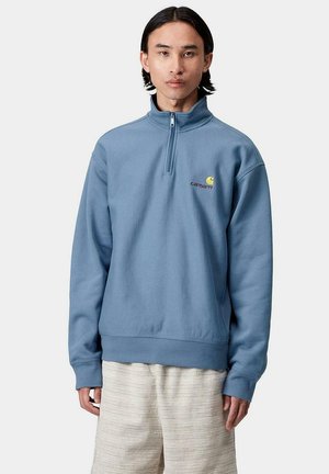 Sudadera ligera de color azul claro con cremallera de un cuarto, hecha de tela suave, que presenta un pequeño logotipo en rojo y amarillo en el lado izquierdo del pecho, con puños y dobladillo acanalados.