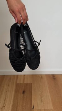 Una mano tiene un paio di ballerine nere in rete con cinturini alla caviglia e piccoli fiocchi, sopra un pavimento di legno e uno sfondo a parete semplice.