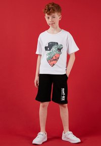 Garçon aux cheveux roux portant un t-shirt blanc avec un motif de dinosaure et un short noir avec le texte "Giant T-Rex", debout devant un fond rouge.