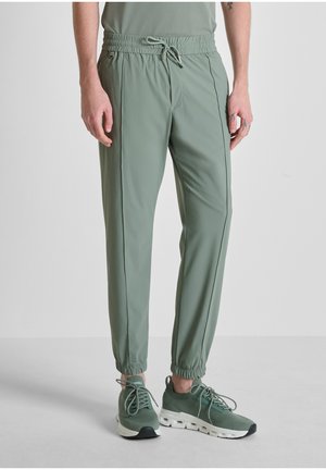 Uomo che indossa pantaloni da jogging verde chiaro e sneakers abbinate, in piedi contro uno sfondo bianco neutro.