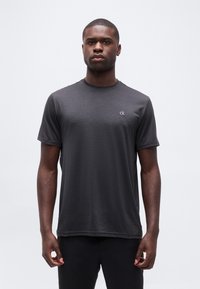 Άνδρας στέκεται προς τα εμπρός, φοράει σκούρο γκρι κοντομάνικο πουκάμισο με ένα μικρό λευκό λογότυπο "ck" στο στήθος, μπροστά σε απλό φόντο.