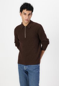 Pull en maille côtelée marron avec un col et une fermeture éclair demi-lune, porté avec un jean bleu. La texture du tissu est douce et la coupe est décontractée.