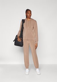 Felpa con cappuccio beige con zip e pantaloni jogger abbinati in materiale morbido, abbinati a una borsa da viaggio nera e scarpe da ginnastica bianche.