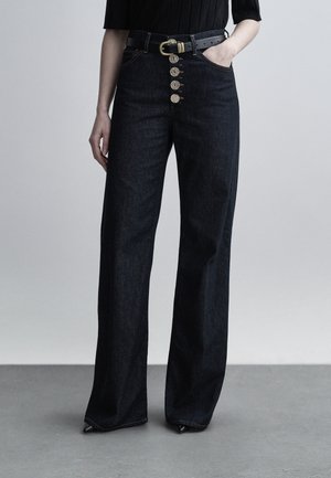 Moteris dėvi juodus plačios kojų formos džinsus su penkiais matomais žalvariniais mygtukais ir juodą diržą su auksine sagtimi, stovinti priešais paprastą foną.