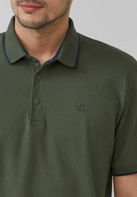 Olivgrünes Poloshirt aus strukturiertem Stoff, mit zwei Knöpfen am Kragen und marineblauen Akzenten am Kragen. Mit kleinem Logo auf der Brust.