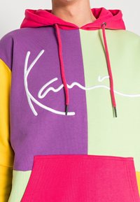 Sudadera colorida con paneles morados, verdes, amarillos y rosas. Tiene un logotipo de firma en blanco y cordones rosa. Tejido suave y texturizado.