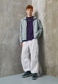 Lichtblauwe zip-up hoodie over een paarse t-shirt, gecombineerd met losse witte cargobroeken en zwart/groene sneakers. Betonnen muur als achtergrond.