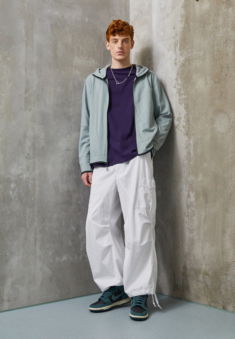 Un sweat à capuche bleu clair zippé sur un t-shirt violet, associé à un pantalon cargo blanc ample et des baskets noir/vert. Fond en mur de béton.