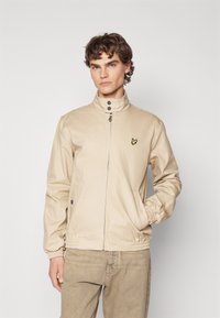 Beige zip-jakke med høy krage, elastiske mansjetter og sidelommer. Har en liten gul logo på brystet. Glatt tekstur.