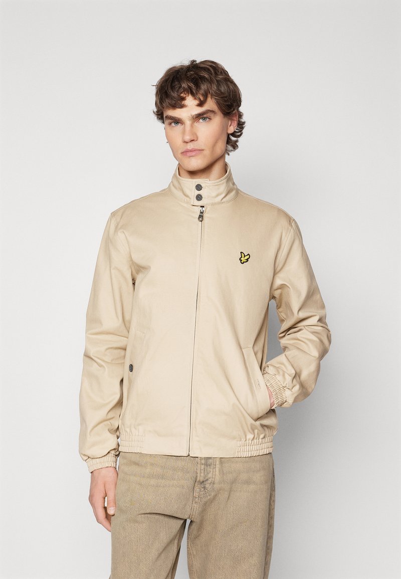 Beige zip-jakke med høy krage, elastiske mansjetter og sidelommer. Har en liten gul logo på brystet. Glatt tekstur.