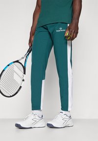Teal træningsbukser med hvide side-striber, featuring et logo på låret og elastiske manchetter. Båret med hvide sneakers og holder en tennisketsjer.