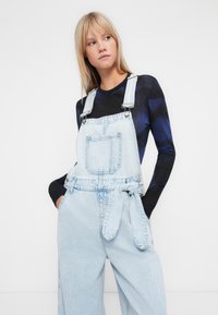 Lichtblauwe denim overall met een losse pasvorm, verstelbare bandjes, een voorkantzak en een strikdetail in de taille, gecombineerd met een donkere top met lange mouwen.