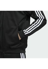 Svart zip-up jacka i jämn tyg, med vita tre-stripes detaljer på ärmarna. Inkluderar sidofickor och ribbade ärmslut.