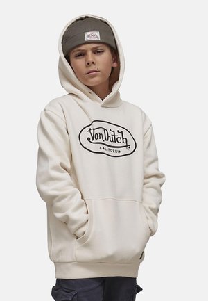 Garçon portant un sweat à capuche beige Von Dutch et un bonnet olive, les mains dans les poches du sweat, regardant directement la caméra avec une expression neutre.