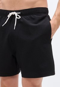 Μαύρο μαγιό με υφή και ραβδωτό σχέδιο, ελαστική μέση και λευκό κορδόνι. Ορατές οι πλευρικές τσέπες, το μήκος κόβεται πάνω από το γόνατο.