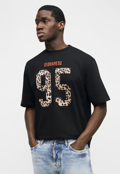 Jeune homme portant un T-shirt noir avec l'inscription « DSQUARED2 » et un numéro « 95 » imprimé en léopard, associé à un jean bleu clair, les mains dans les poches, faisant face à gauche.
