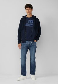 Granatowa kurtka z kapturem na zamek z prążkowanej tkaniny, noszona na granatowej koszulce z grafiką. Połączona z jasnoniebieskimi dżinsami i białymi trampkami.