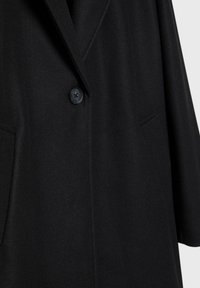 Zwarte wollen jas met een enkele zwarte knoop, gelapte kraag en twee zijzakken, weergegeven in close-up.