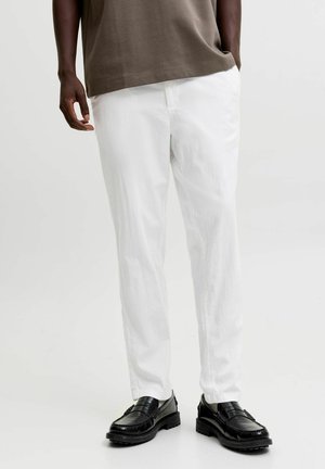 Personne à la peau foncée portant un pantalon blanc, une chemise marron et des mocassins en cuir noir, debout avec une main dans la poche.