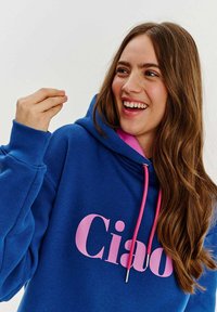 Modrá mikina s růžovým nápisem "Ciao", se šňůrkami a texturovanou tkaninou. Dlouhé rukávy a volný střih, nošena usměvavou osobou.