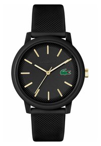 Montre ronde noire avec bracelet texturé noir, marqueurs d'heure et aiguilles dorés. Présente le logo Lacoste en vert sur le cadran. Mouvement à quartz.