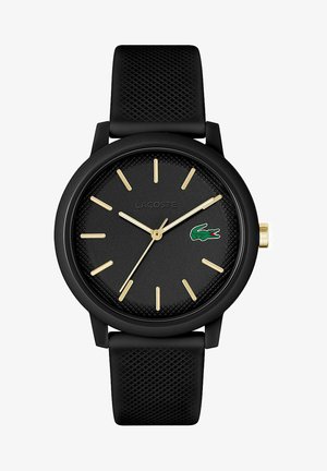 Montre ronde noire avec bracelet texturé noir, marqueurs d'heure et aiguilles dorés. Présente le logo Lacoste en vert sur le cadran. Mouvement à quartz.
