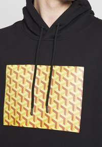 Black hoodie featuring a rectangular yellow graphic with a repeating 3D triangle pattern in orange and red tones. Black drawstrings included.