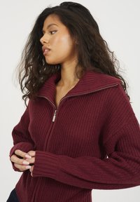 Burgundowy sweter z ribbowanym splotem, z wysokim kołnierzem, pełnym zamkiem błyskawicznym i szerokimi rękawami. Miękka tekstura z wyraźnym wzorem dzianiny i dopasowanymi mankietami.