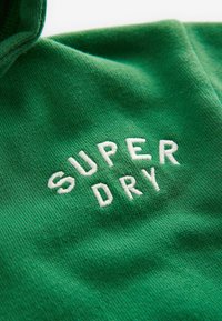 Zeleni dukserica s oznakom "SUPER DRY" u bijelom vezenom tekstu. Tkanina ima mekanu teksturu i udoban, opušteni kroj.