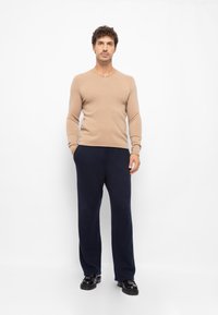 Beige Strickpullover mit V-Ausschnitt, kombiniert mit navyfarbenen, weit geschnittenen Hosen und schwarzen Lederschuhen. Glatte Stoffstruktur, figurbetonte Silhouette.