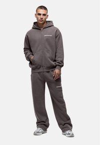 Grijze zip-up hoodie met kangoeroezakken, gecombineerd met bijpassende grijze joggingsbroek en lichte sportschoenen. Heeft logo-tekst in het wit.