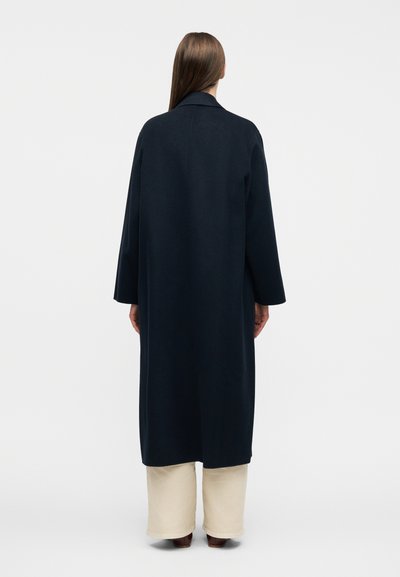 Γυναίκα με μακριά καστανά μαλλιά, ντυμένη με μακρύ σκούρο ναυτικό παλτό, εκρού παντελόνι με φαρδιά μπατζάκια και καφέ παπούτσια, στέκεται με την πλάτη γυρισμένη σε ένα απλό λευκό φόντο.