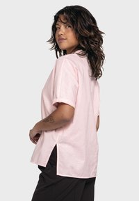 Hellrosa Kurzarmshirt mit locker sitzendem Schnitt, Seitenschlitzen und glatter Textur. Einfaches Design, klassischer Kragen und umgeschlagene Ärmel.