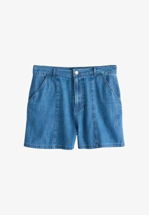 Femeie purtând o cămașă albă cu nasturi și mâneci tubulare și pantaloni scurți din denim albastru cu talie înaltă, cu mâinile în buzunare, trunchiul și partea superioară a picioarelor vizibile.