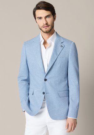 Mężczyzna z krótkimi brązowymi włosami i brodą, ubrany w jasnoniebieską marynarkę na białej koszuli i białe spodnie, stojący na jednolitym tle.