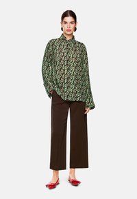 Grün und schwarz gemusterte Bluse mit Kragen, lockerer Passform und langen Ärmeln, kombiniert mit braunen, weit geschnittenen Hosen und roten Ballerinas.