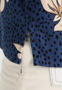 Blouse à motifs bleus avec grands imprimés floraux et pois noirs. Tissu léger avec une fente latérale et une texture lisse.
