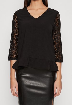 Kobieta ubrana w czarną bluzkę z dekoltem w serek, trzy czwarte rękawa z koronką i marszczonym obszyciem, w połączeniu z czarną skórzaną ołówkową spódnicą.