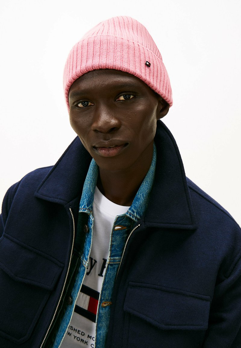 Różowa dzianinowa czapka, granatowa wełniana kurtka i dżinsowa koszula nałożona na białą koszulkę z graficznym detalem. Zbliżenie na twarz modela.