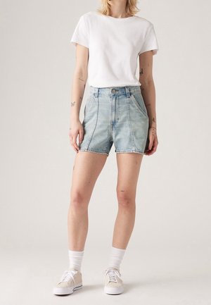 Osoba ubrana w gładki biały t-shirt, jasnoniebieskie dżinsowe szorty z wysokim stanem, białe skarpetki i beżowe tenisówki, stojąca na jednolitym tle.