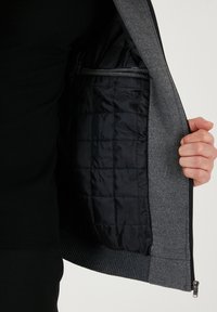 Sivá zipsová bunda s textúrovaným vonkajším povrchom a čiernym polstrovaným vnútrom, ktoré predvádza prešívaný vzor a rebrované manžety.