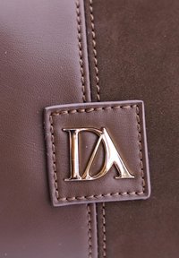 Material de cuero marrón con un acabado suave, que presenta una placa de logo dorada y costuras detalladas a lo largo de los bordes.