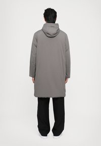 Un long manteau gris avec une capuche, présentant un design simple et une texture matelassée. Un pantalon noir et des chaussures blanches complètent la tenue.