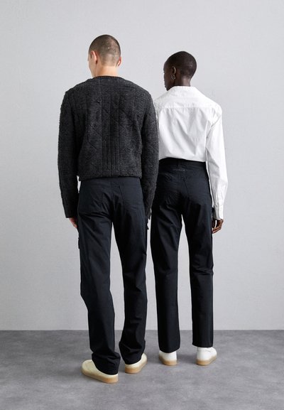 Black trousers on two figures, one wearing a textured dark sweater, the other a white shirt. Both wear light-colored casual shoes.