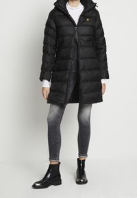 Henkilö, jolla on yllään musta huppupäinen puffatakki valkoisen paidan, harmaiden kapeiden farkkujen ja mustien nilkkabootsien kanssa, seisoo yksinkertaista valkoista taustaa vasten.