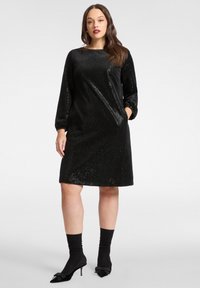 Abito nero con paillettes, maniche lunghe, vestibilità rilassata, scollo rotondo e tasche laterali. Indossato con calzini neri al ginocchio e tacchi a punta.