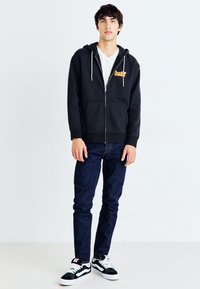 Sudadera negra con cremallera y un bolsillo frontal, con cuerdas de ajuste blancas; llevada sobre una camiseta blanca, combinada con jeans oscuros y zapatillas negras.