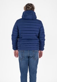 Man in een blauwe gewatteerde jas met een capuchon en blauwe jeans, staande met zijn rug naar de camera tegen een witte achtergrond.