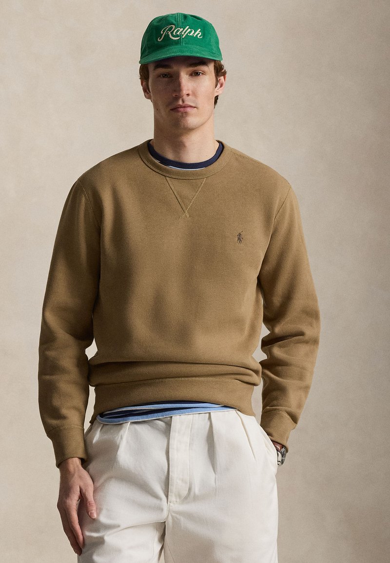 Νεαρός άνδρας που φορά πράσινο καπ έλο με το κείμενο «Ralph», γκριζωπό φούτερ, φορεμένα πουκάμισα σε στρώσεις και λευκό παντελόνι με πιέτες, στέκεται μπροστά σε ουδέτερο φόντο.